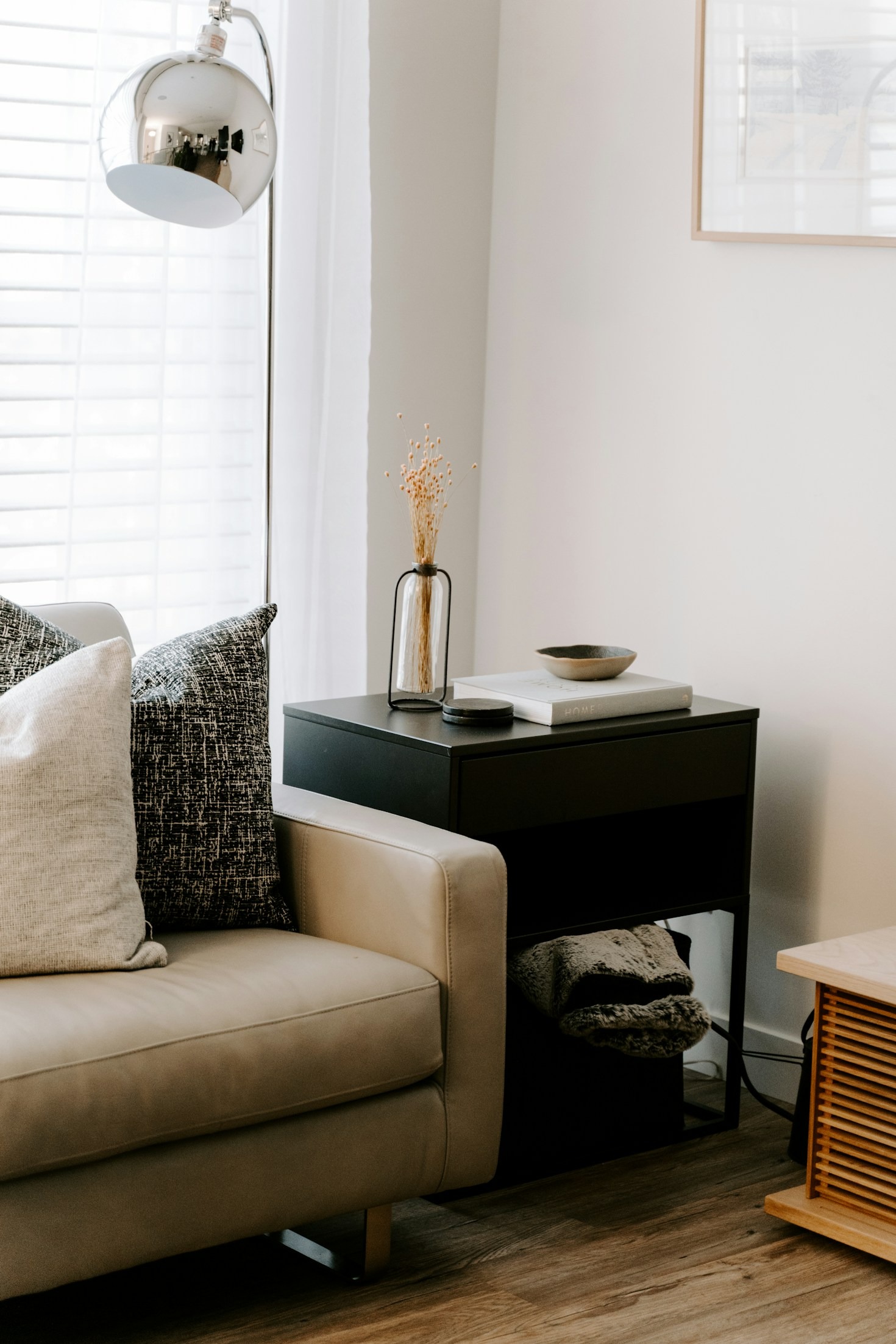 Modern living room with a beige leather sofa, decorative pillows, a sleek black side table with minimalist decor, and a silver floor lamp near a window with white blinds